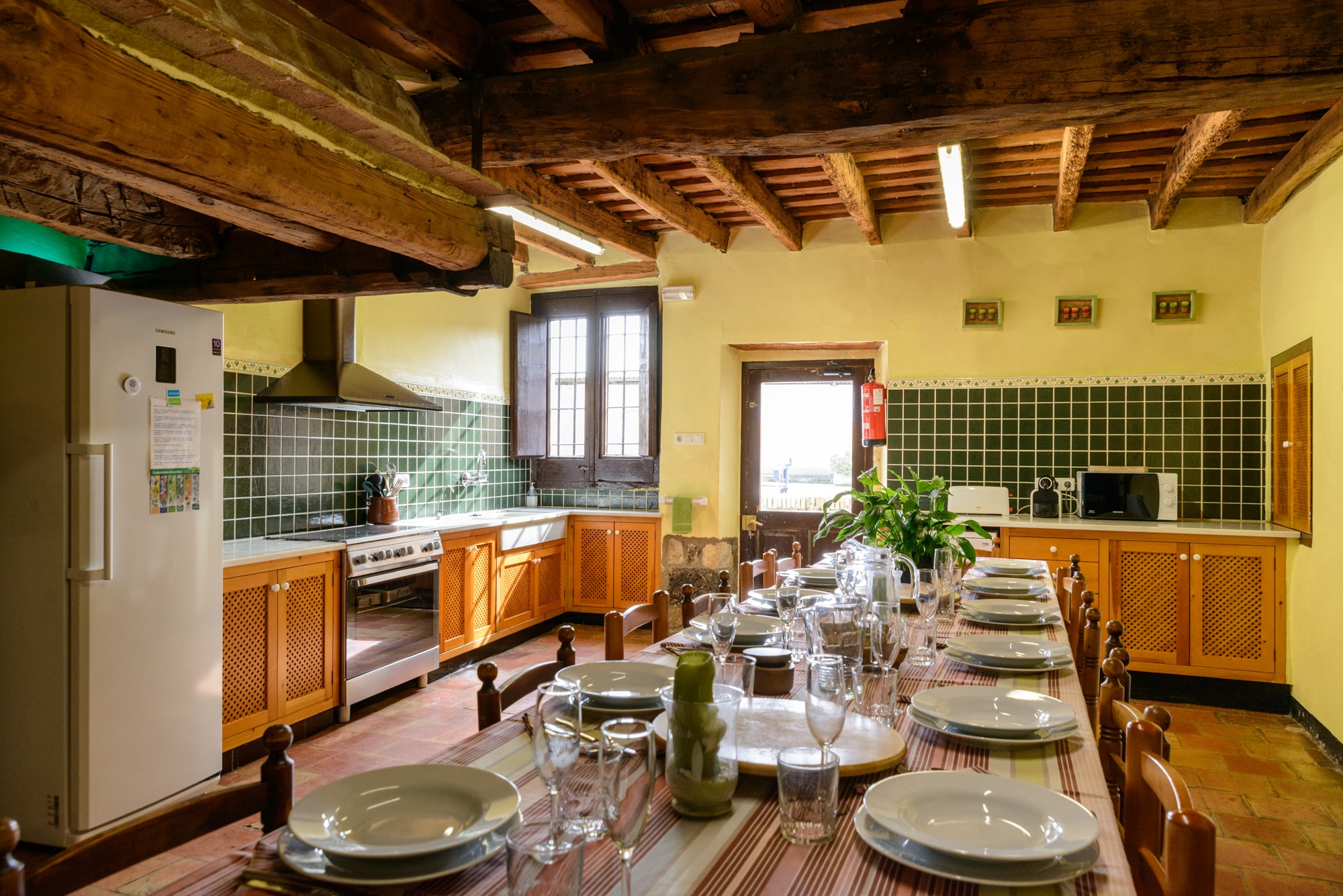 Cocina grande con la mesa preparada, la mesa es larga y caben unos 14 comensales. Techo con vigas de madera y las paredes tienen azulejos verdes.