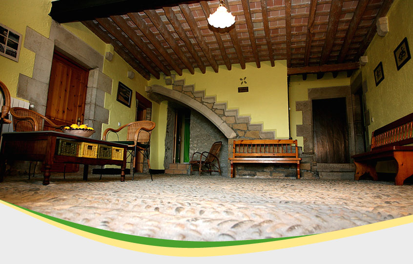 Hall de entrada de la casa con dos bancos de madera para sentarse, una mesita pequeña de madera y alrededor 3 sillas. Tiene un espacio vacio grande en medio de la sala con varias puertas por alrededor para acceder a las demás habitaciones.
